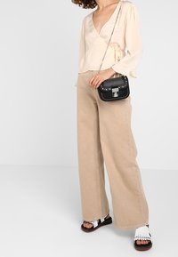 Kvinna klädd i en beige omslagstopp, vida tan byxor, vita franssandaler och håller en liten svart spikes crossbody-väska.