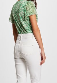 Top verde con motivo floreale e maniche corte arricciate, abbinato a jeans bianchi attillati con tasche posteriori e dettaglio con bottone.