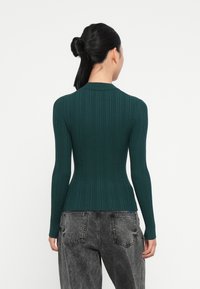 Rippstrick, figurbetontes grünes Sweatshirt mit langen Ärmeln und Kragen. Die Rückansicht zeigt die Textur und Konturen an der Taille. Kombiniert mit hochsitzzenden Jeans.