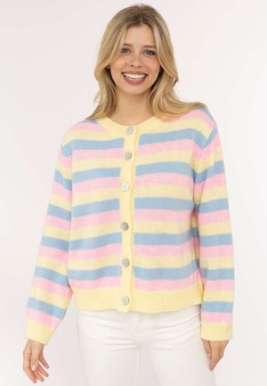 Frau lächelt und trägt eine pastellgelbe, rosa und blaue gestreifte Knopfleisten-Strickjacke mit weißen Hosen vor einem schlichten Hintergrund.
