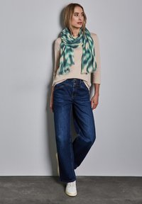 Maglietta beige a maniche lunghe, jeans blu a gamba larga e una sciarpa a motivo teal. La sciarpa ha un design texturizzato e bordi frangiati. Sneakers bianche completano il look.