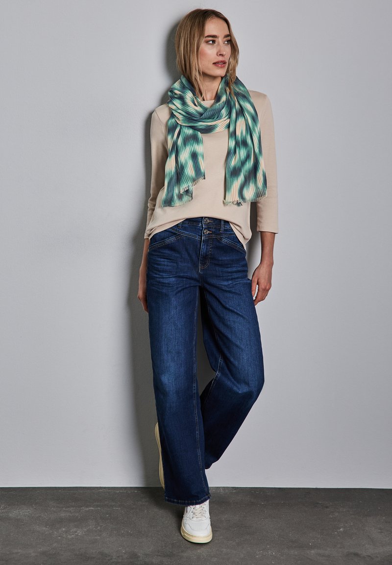 Maglietta beige a maniche lunghe, jeans blu a gamba larga e una sciarpa a motivo teal. La sciarpa ha un design texturizzato e bordi frangiati. Sneakers bianche completano il look.