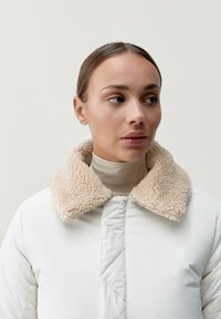 Biała kurtka puchowa z beżowym kołnierzem z owczej wełny, z pikowaną fakturą i zapięciem na zamek, noszona na jasnobeżowym golfie.