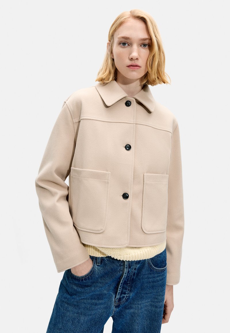 Młoda kobieta o blond włosach, ubrana w beżową zapinaną na guziki kurtkę na kremowym swetrze i niebieskich dżinsach, stojąca z jedną ręką w kieszeni.