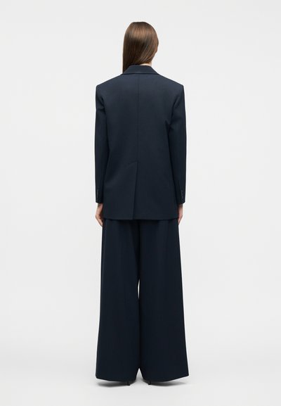 Γυναίκα με μακριά ίσια μαλλιά, φορώντας ένα σκούρο μπλε κομψό σακάκι και φαρδιά παντελόνια, όρθια με την πλάτη στραμμένη προς ένα απλό φόντο.