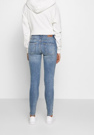 Person iført lyseblå skinny jeans, hvid hættetrøje og hvide sneakers, stående vendt mod en ensfarvet hvid væg.