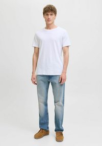 Joven de pie vistiendo una camiseta blanca lisa, jeans azul claros y zapatos de ante marrones frente a un fondo neutro.