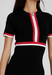 Robe tricot à manches courtes noire avec des bordures rayées rouges et blanches sur les manches, la taille et une fermeture éclair à l'avant allant du col au milieu de la poitrine.
