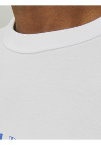 Hvid T-shirt med rund halsudskæring, korte ærmer og et tekstureret bomuldsstof med blå grafisk tekstdetalje nær kanten.