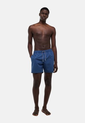 Jeune homme debout pieds nus, portant un short de bain bleu avec cordon, une main dans la poche, sur un fond clair uni.