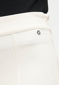 Κοντινό πλάνο ενός ατόμου που φοράει λευκά παντελόνια ψηλής μέσης με εμφανές ασημένιο λογότυπο "G" στο πλάι και μαύρο τοπ στερεωμένο μέσα στο παντελόνι.