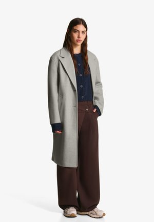 Grå uldfrakke med struktureret design, blå strik cardigan under, brune brede bukser og beige sneakers.