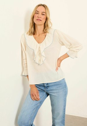 Cream checkered blouse with ruffled V-neck, light blue scalloped trim, and three-quarter sleeves, paired with light blue jeans.