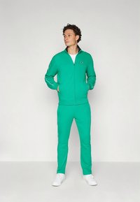 Zaļš rāvējslēdzēja tracksuit no mīksta auduma, ar baltām detaļām uz piedurknēm un sānos, kopā ar baltām sporta kurpēm.