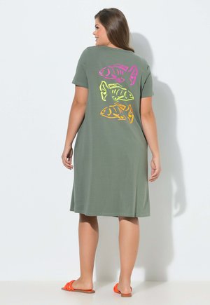 Frau im olivgrünen Kleid mit drei farbenfrohen Fischmotiven auf dem Rücken steht in orangefarbenen Sandalen vor weißem Hintergrund.