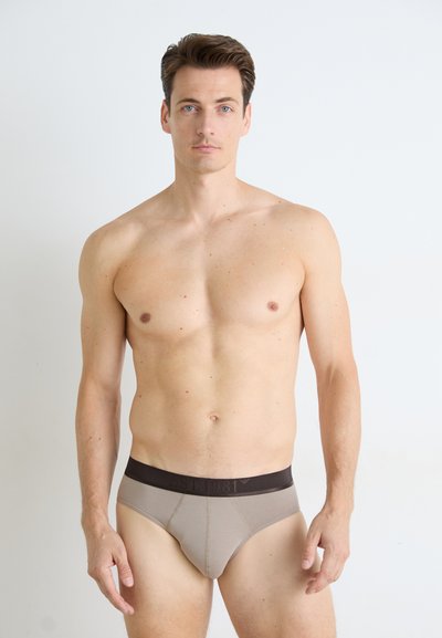 Beige bomullsboxershorts med mørkebrunt liv, som har preget logo, tettsittende design og glatt tekstur. Modellen står med ansiktet fremover.