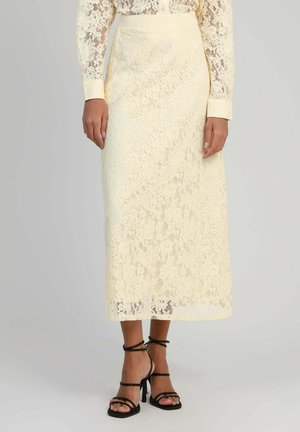 Femme portant une jupe midi en dentelle crème assortie à un chemisier en dentelle et des sandales à talons noires à lanières, debout devant un fond uni.