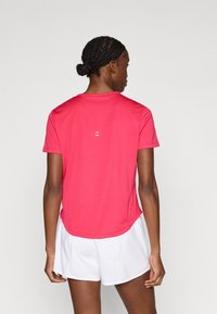 Αθλητική μπλούζα με κοντά μανίκια σε έντονο ροζ χρώμα, φτιαγμένη από λείο ύφασμα. Διαθέτει στρογγυλεμένο τελείωμα και μικρό λευκό λογότυπο στο πίσω μέρος. Συνδυάζεται με λευκά σορτς.