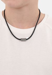 Collar de cordón trenzado negro con un colgante cilíndrico de plata texturizada. Lleva puesto sobre una camisa blanca, mostrando el detalle y diseño del colgante.