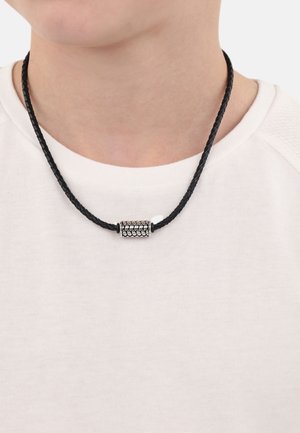 Collana in corda intrecciata nera con un pendente cilindrico textured in argento. Indossata su una camicia bianca, mette in risalto i dettagli e il design del pendente.