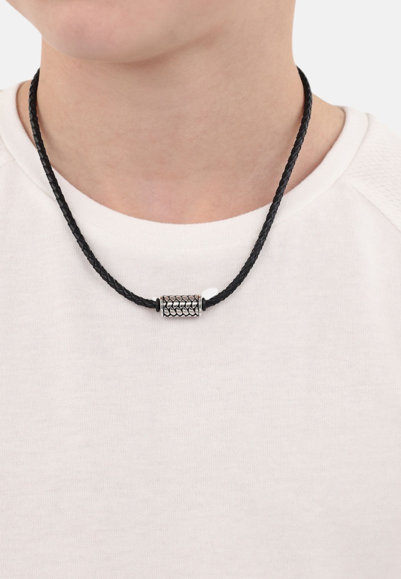 Collar de cordón trenzado negro con un colgante cilíndrico de plata texturizada. Lleva puesto sobre una camisa blanca, mostrando el detalle y diseño del colgante.