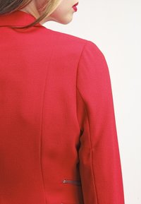 Blazer rouge en tissu texturé, coupe cintrée, col cranté, et un accent de fermeture éclair argentée sur le côté gauche. Design classique et structuré.