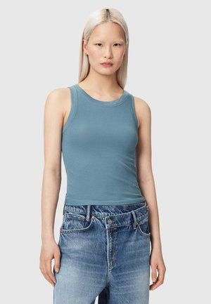 Jonge vrouw met steil platina blond haar draagt een aansluitend blauwe mouwloze top en hoge losse blauwe spijkerbroek tegen een effen achtergrond.