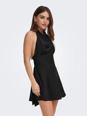 Jeune femme portant une robe dos nu en satin noir, des boucles d'oreilles créoles dorées et un bracelet en or, debout devant un fond clair uni.