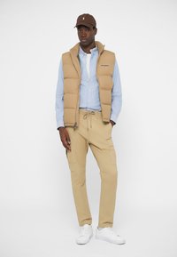 Tan puffer vest over een lichtblauw overhemd met knopen, gecombineerd met tan cargobroeken en witte sneakers. Een bruine pet voegt een contrasterend detail toe.