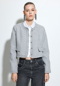 Chaqueta de tweed gris recortada con cuello redondo, bolsillos delanteros y cierre de botón. Lleva puesta sobre una camisa blanca y combinada con jeans oscuros.