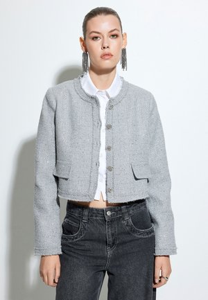 Giacca cropped in tweed grigio con scollatura rotonda, tasche frontali e chiusura con bottoni. Indossata sopra una camicia bianca e abbinata a jeans scuri.