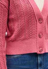 Ροζ πλεκτή ζακέτα με υφασμένο σχέδιο σε σχήμα ζικ-ζακ, μεγάλα στρογγυλά κουμπιά, φορεμένη πάνω από λευκό ριμπ μπλουζάκι και μπλε τζιν.