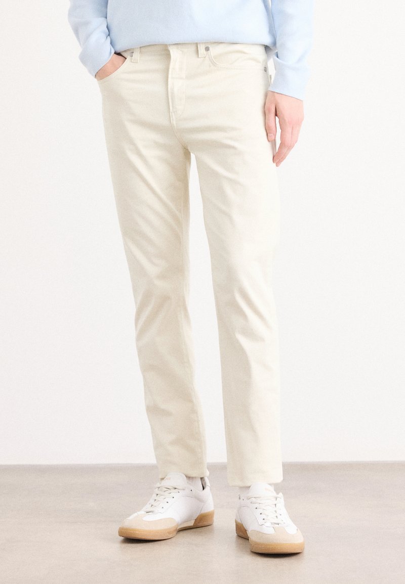 Lichtbeige slim-fit broeken met vijf zakken, subtiele textuur en een rechte pijp, gecombineerd met witte sneakers met rubberen zolen.