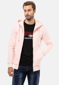 Lichtroze zip-up hoodie van zacht materiaal. Bevat zakken aan de voorkant en een capuchon met koord. Gelaagd over een zwart graphic t-shirt en jeans.
