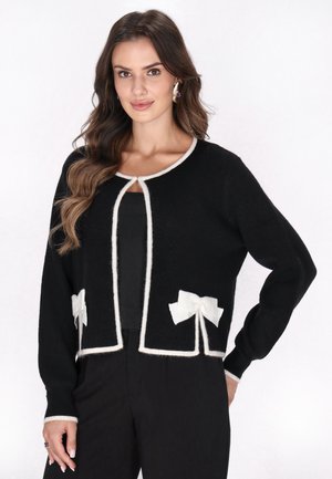Schwarze Strickjacke mit tailliertem Schnitt, weißem Saum und zwei weißen Schleifenakzenten an den Seiten. Weicher Stoff mit langen Ärmeln und offenem Vorderteil.