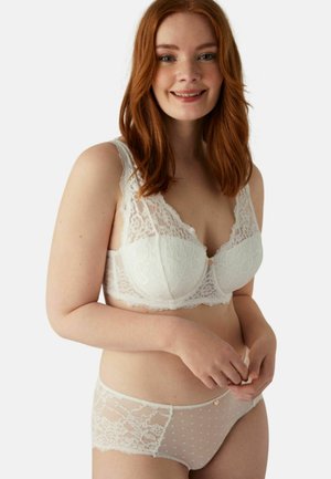 Sujetador de encaje blanco con borde festoneado y detalle central con botón, combinado con bragas transparentes de lunares que presentan acentos de encaje en los laterales.