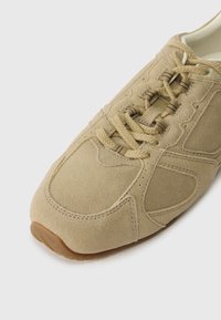 Sapatilha de camurça bege com bico arredondado, painéis texturizados, atacadores a condizer e sola de borracha. Detalhes cosidos em volta das bordas.