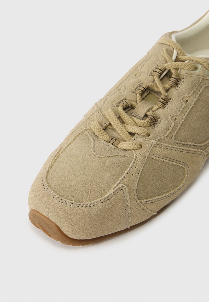Beige suede sneaker with a rounded toe, textured panels, matching laces, and a rubber outsole. Stitched detailing around the edges.