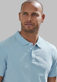 Polo bleu clair en tissu texturé, avec un col pointu, un patte de boutonnage à trois boutons et un petit logo brodé sur la poitrine.