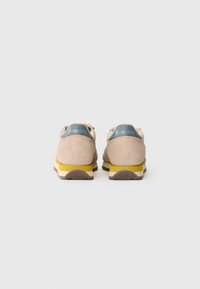 Sneaker in suede beige con suola gialla e crema, caratterizzati da un logo blu sul retro e un design minimalista.
