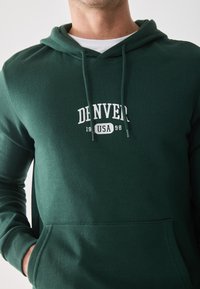 Groene hoodie van zacht materiaal met een witte opdruk "DENVER 1998 USA" op de voorkant, met een capuchon met trekkoord en een voorzak.