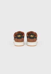 Hnedé semišové tenisky s čiernymi akcentmi, s bielou gumovou podrážkou a žltým logom na päte. Zaoblený tvar, nízky profilový dizajn.