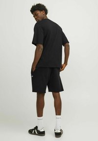 Juodas medvilninis t-shirt ir šortų komplektas su laisvu pritaikymu. Matomos šoninės kišenės šortuose. Modelis nešioja juodus sportbačius ir baltas kojines.