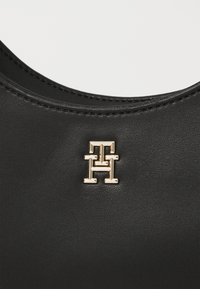 Zwarte synthetische handtas met een matte afwerking, voorzien van een gouden logoaccent. De gladde textuur en ronde hoeken versterken het moderne ontwerp.