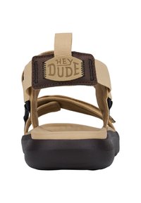 Beżowe sandały z brązową skórzaną naszywką oznaczoną "HEY DUDE". Posiadają regulowane paski oraz teksturowaną brązową podeszwę dla lepszej przyczepności.