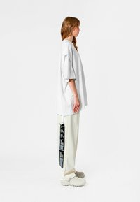 Camiseta blanca de gran tamaño con hombros caídos, combinada con pantalones crema que muestran un detalle de etiqueta negra y calzado blanco texturizado.