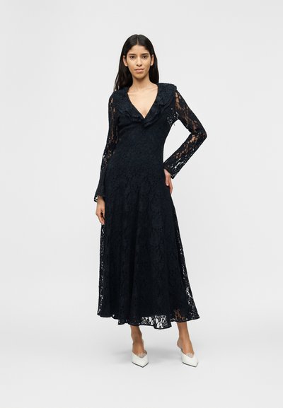 Γυναίκα με μαύρο midi φόρεμα από δαντέλα με μακριά μανίκια και λαιμόκοψη με βολάν, όρθια με το ένα χέρι στη μέση, φοράει λευκά μυτερά τακούνια.