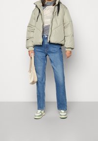 Gewatteerd jack in lichtgrijs, gecombineerd met een patroontrui, blauwe hooggetailleerde spijkerbroek en groene en witte sneakers. Kleine beige tas.