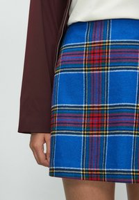 Blau karierten Rock mit einer wollartigen Textur, der rote, grüne und gelbe Akzente aufweist, kombiniert mit einer kastanienbraunen Jacke im Vordergrund.