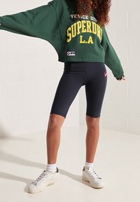 Person wearing green cropped Superdry sweatshirt, black cycling shorts, white socks, and white sneakers standing against white wall.